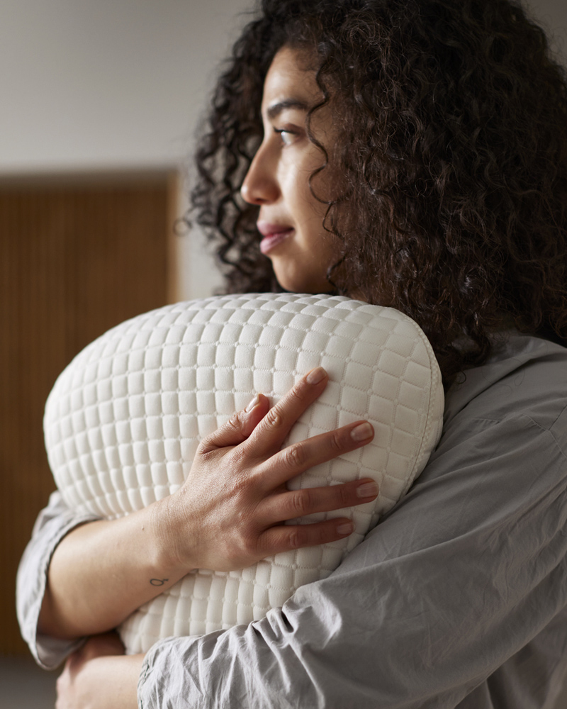A person holding a KLUBBSPORRE ergonomic pillow for side and back sleepers to their chest.