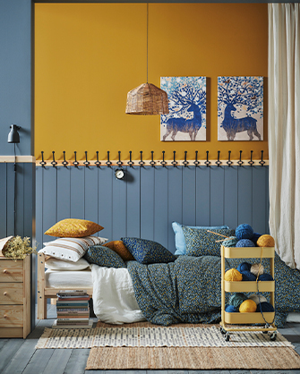 Dormitorio con pared bicolor (mostaza y azul panelado con ganchos) y cama de madera con cojines estampados y textiles azules, flanqueada por una mesita de noche de madera y un carrito con lanas de colores, y decorado con dos cuadros de ciervos.