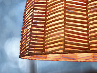 Detalle de una lámpara colgante de pantalla de bambú tejido con un patrón de espiga, mostrando cómo la luz se filtra a través de las aberturas creando un efecto cálido y texturizado.