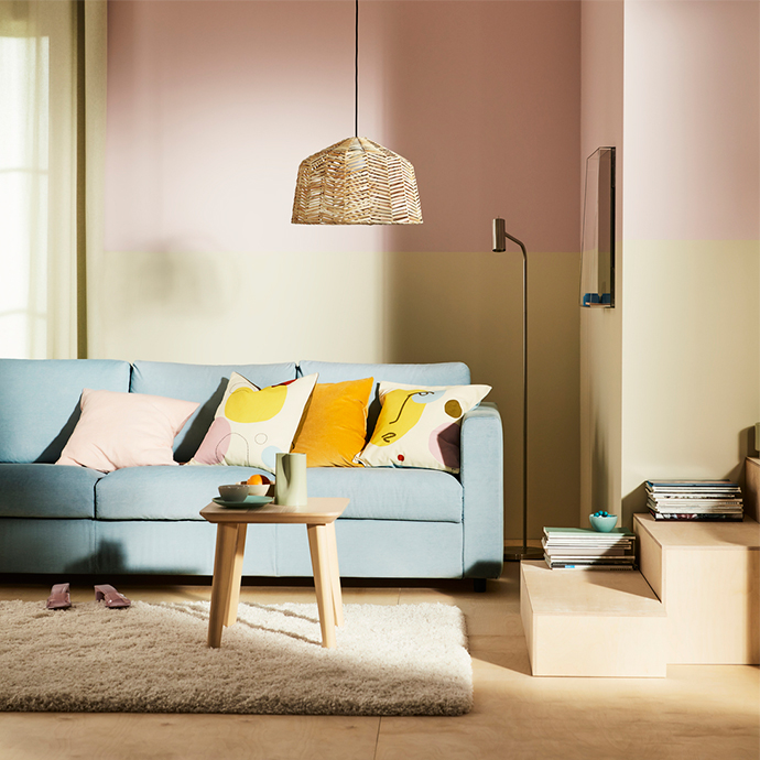 Salón con paredes de dos tonos (rosa y beige), un sofá azul claro con cojines decorativos de colores pastel y amarillos, una mesa de centro de madera clara, una alfombra mullida beige y una lámpara de techo de mimbre.