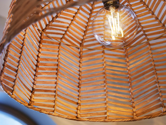 Primer plano de una lámpara colgante de pantalla de bambú tejido con un patrón geométrico, revelando una bombilla de filamento vintage encendida en su interior.