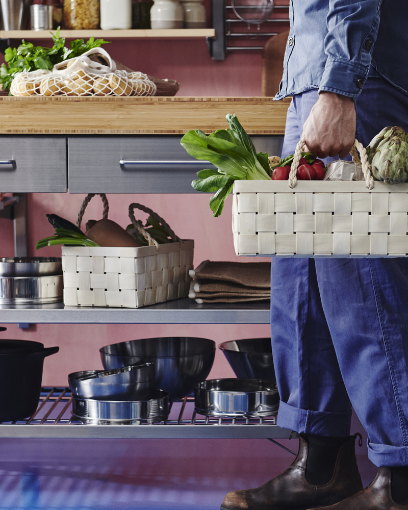 A person holds a poplar KÖSSEBÄR harvesting basket filled with vegetables, standing by a work bench with shelves and drawers.