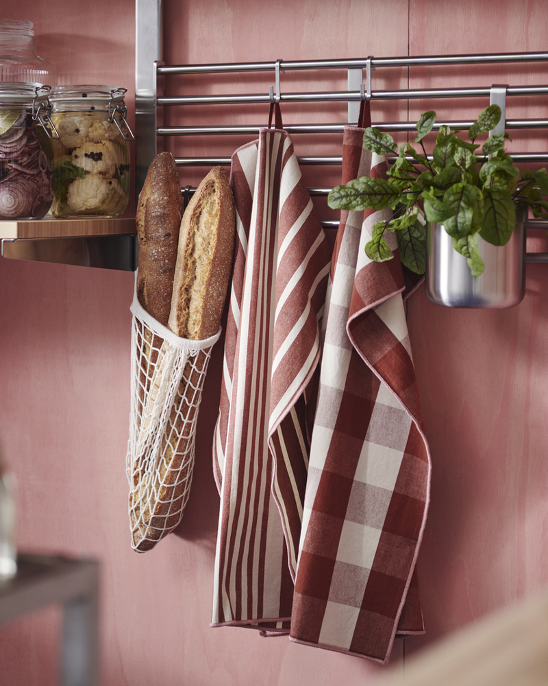 Two KÖSSEBÄR tea towels with mixed patterns, a mesh bag with bread, and a potted plant are hung on the rails of a wall shelf.
