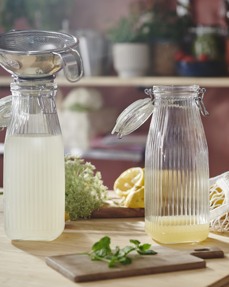 Two KÖSSEBÄR bottle shaped jars with lids, part-filled with yellowish liquid, sit on a worktop. One jar has a funnel in it. 