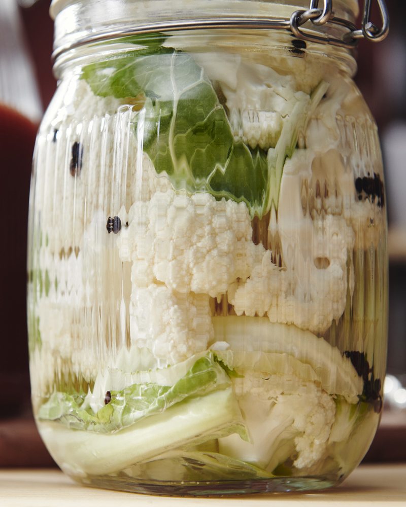 A clear glass KÖSSEBÄR jar with lid, filled with cauliflower and other vegetables, sits on a wooden worktop. 