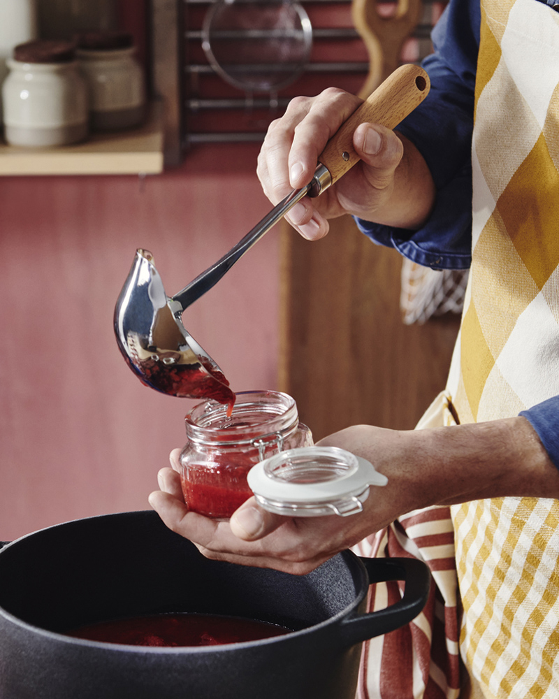 A person wearing an apron holds a VARDAGEN soup ladle in one hand and a glass jar in the other, spooning jam into the jar.