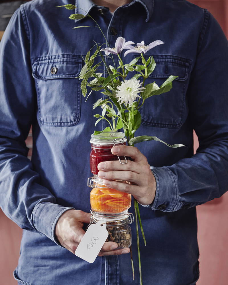 A person is holding some flowers and three KÖSSEBÄR jars with lids filled with different foods in front of themselves.
