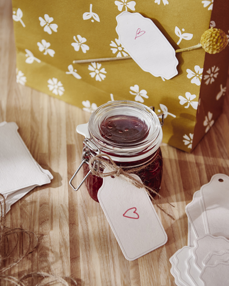 A glass jar with a lid filled with jam sits on a worktop. A label with a written greeting is attached to the jar by a string.