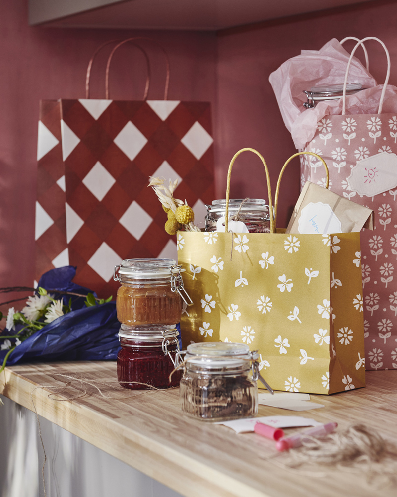 Three KÖSSEBÄR gift bags in mixed patterns sit on a worktop beside a bouquet of flowers and several jars filled with foods. 