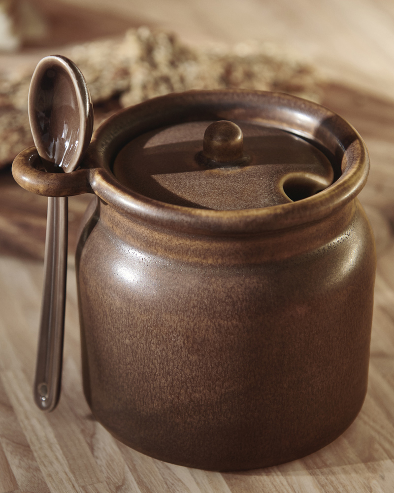 A brown KÖSSEBÄR jar with spoon sits on a wooden surface. The spoon is neatly placed in the loop on the side of the jar.