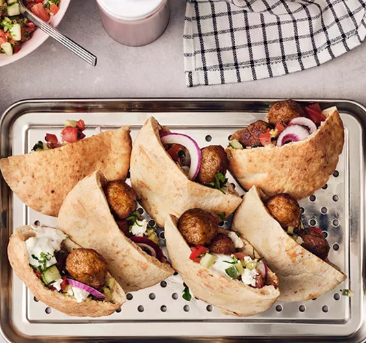 Opened pita breads filled with meatballs, red onion, cucumber, tomato, and creamy white sauce, presented on a perforated metal tray.