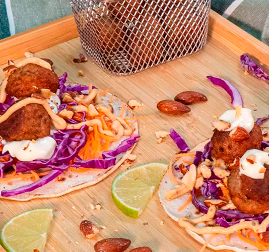 Meatball tacos served on a wooden platter, topped with shredded purple cabbage, grated carrots, white sauce, toasted almonds, and parsley, accompanied by lime wedges and a metal basket with more meatballs.