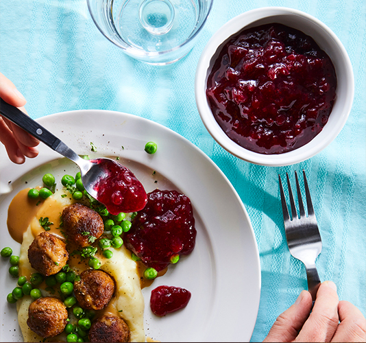 White plate with mashed potatoes, meatballs, and peas, accompanied by cranberry sauce and a glass of water, with a person serving more sauce onto the plate.