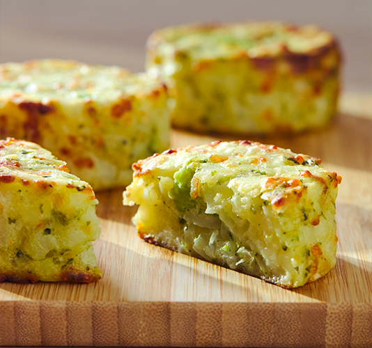 Baked potato and vegetable patties or medallions with gratinated cheese on top, presented on a wooden board, with one of them cut open to show the interior.