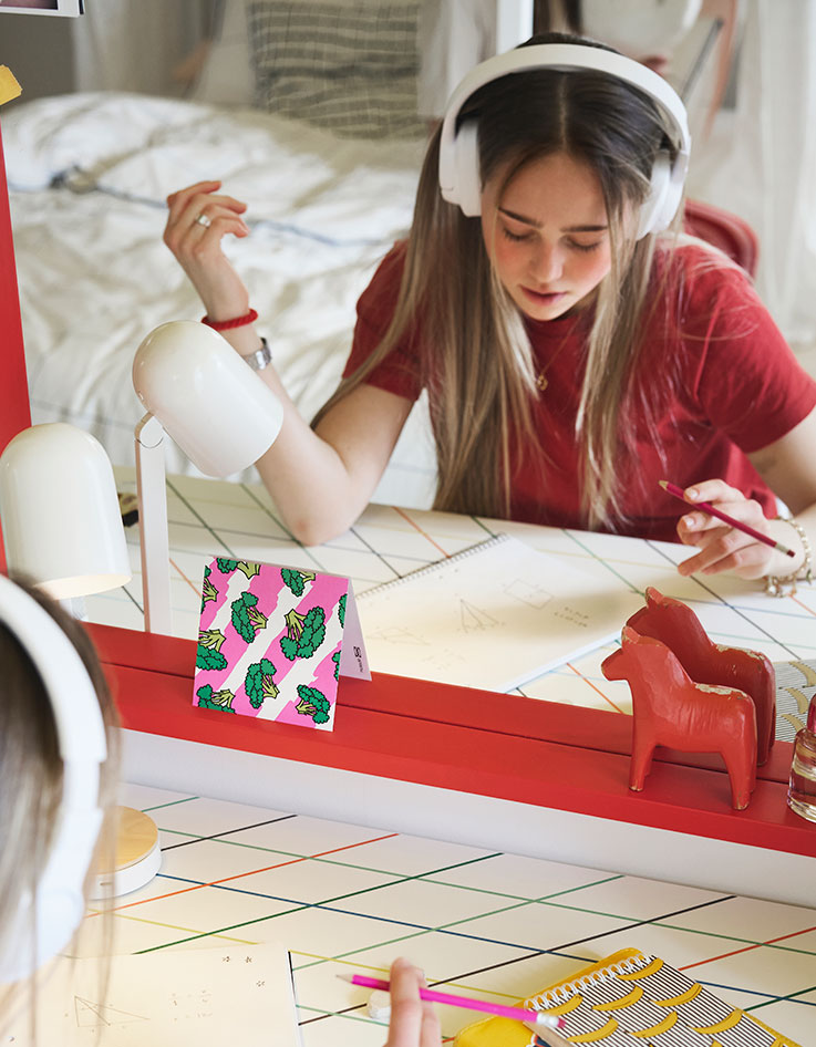 A teenager sitting studying by a LAGKAPTEN/ SPÄND desk with a colourful grid pattern placed in front of a big red mirror.