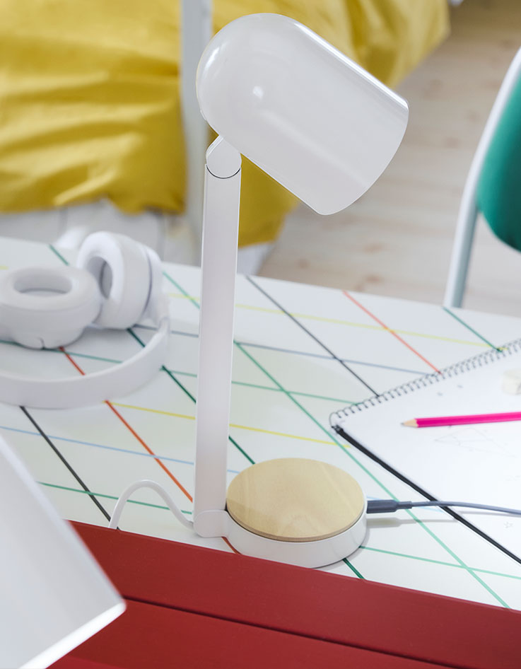 The white ENSMÄRKE desk lamp placed on a desk with a graphic pattern with a bed in the background made with yellow bedlinen