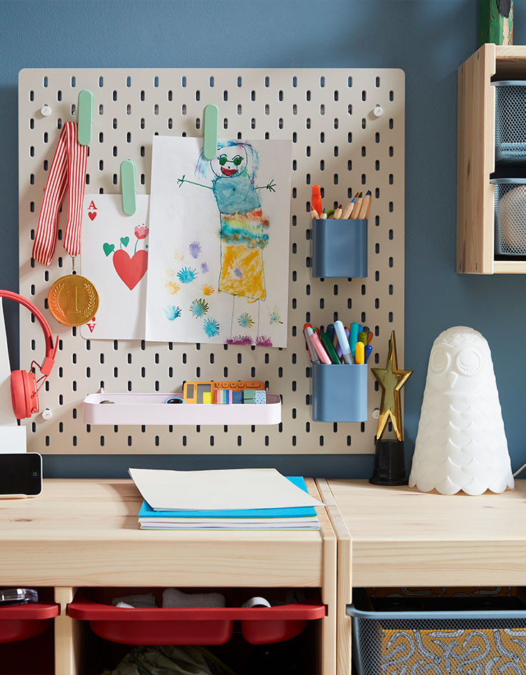 A beige SKÅDIS pegboard with colourful accessories holding pens and drawings placed on a blue wall above storage in pine.