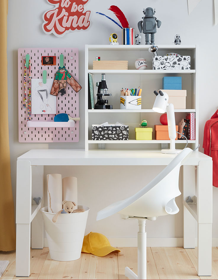 A white PÅHL desk and a white desk chair placed in a children’s room with a shelf and a pink SKÅDIS pegboard