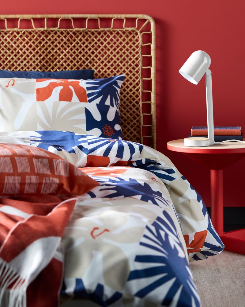 A red bedroom features a bed with a woven headboard and bedding with a blue, orange, and white leaf pattern.