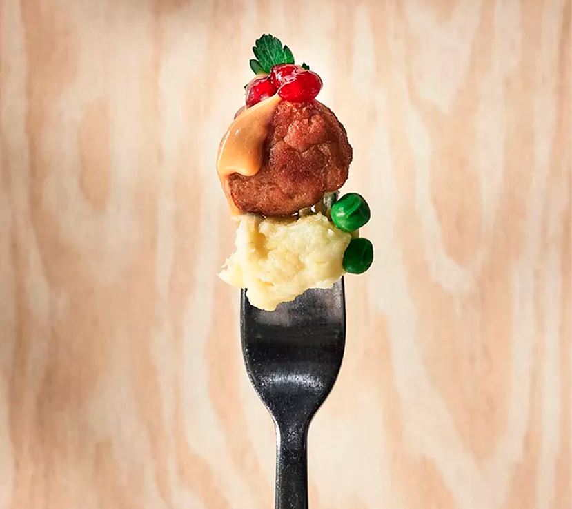 A meatball topped with gravy, cranberries, and a sprig of parsley, stacked on a mound of mashed potatoes and two green peas, all impaled on a fork.
