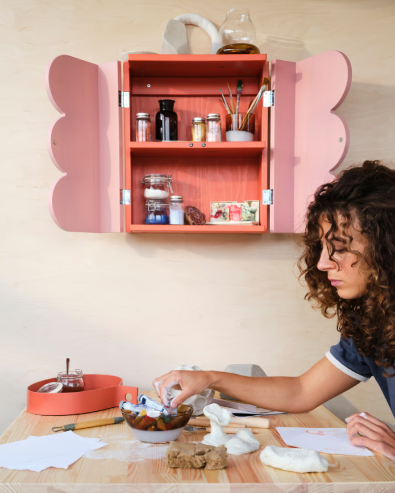 Small rustic cabinet with paintbrushes and paints; girl painting at a table with bowls and sculpting clay