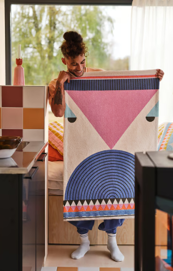 Person holding a colorful blanket with autumnal tones, featuring flat geometric shapes and figures; the blanket appears to be woven.