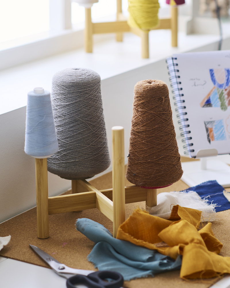 A bamboo DAKSJUS plant stand on top of a white desk holding different coloured rolls of sewing twine, with scissors nearby.