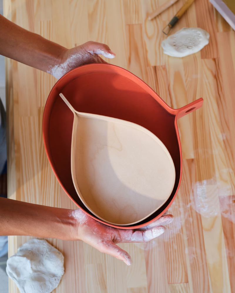 Two hands holding oval wooden trays, the large one and the small one inside the red wood-colored one.