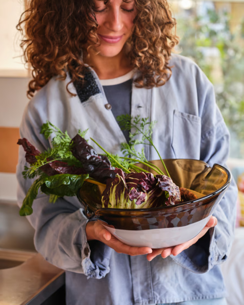 Woman wearing a loose-fitting denim shirt, holding a large dark glass bowl with purple lettuce, carrots, and some herbs.