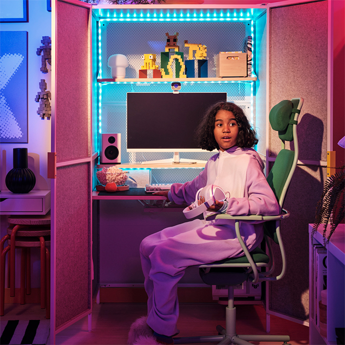 A young person sitting in an gaming chair in a neon-lit gaming corner, wearing purple pajamas, using headphones, and looking at a computer screen.