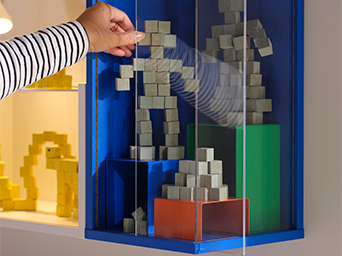 Close-up of a hand placing a piece on a shelf of building blocks, showing block figures in various colors inside a glass display cabinet.