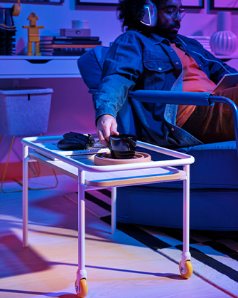A man sitting in a blue armchair, taking a coffee cup from a white rolling serving cart, in a room with neon lighting.