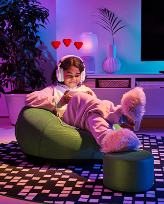 A young person sitting on a green beanbag chair in a living room with neon lighting, playing on a mobile device while wearing white headphones. The scene is decorated with plants and a matching small pouf.