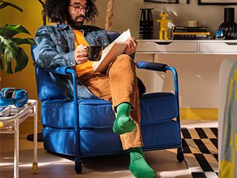 A man with curly hair and a beard, sitting in a blue armchair, reading a book and drinking from a cup in a room with yellow and blue toned walls.