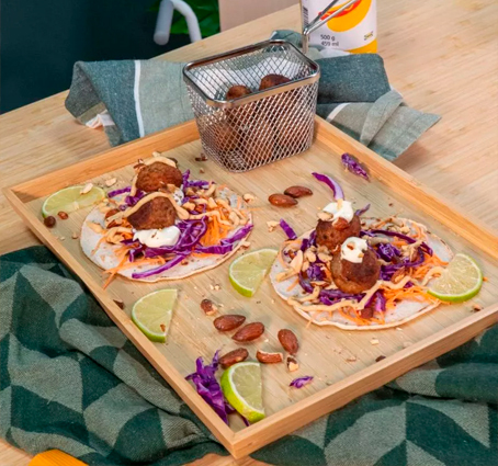 Meatball tacos served on a wooden platter, topped with shredded purple cabbage, grated carrots, white sauce, toasted almonds, and parsley, accompanied by lime wedges and a metal basket with more meatballs.
