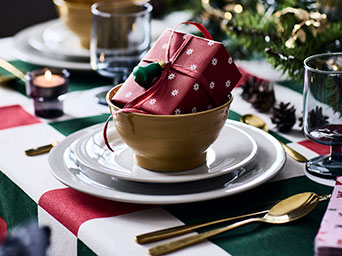A small present sits in a bowl on a festive dinner table, wrapped in a red VINTERFINT gift wrapper with a snowflake pattern.