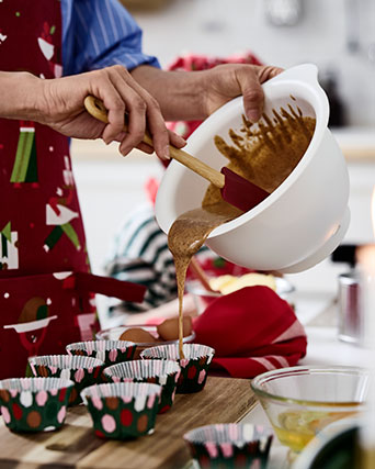A person wearing an apron pours cake mixture onto festive VINTERFINT baking cups, on a cutting board, on a table.