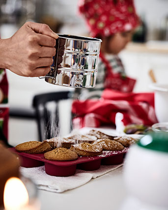 A person wearing an apron is sprinkling powdered sugar over muffins in a muffin mould.