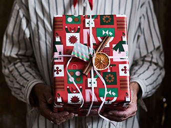 A person with a striped shirt is holding a present wrapped in VINTERFINT gift wrapper and adorned with a dried fruit.
