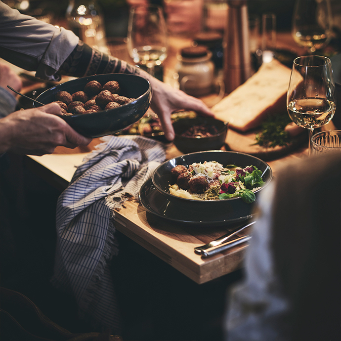 Hands holding a serving dish of matte black stoneware bowls with meatballs, a gray striped napkin, next to a plated meal, a wooden serving board, and a wine glass.
