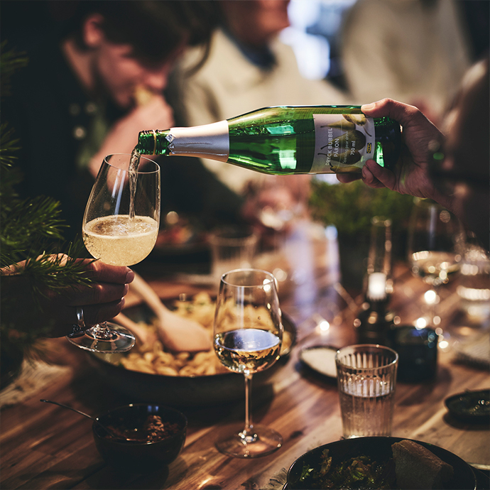 Close-up of a hand pouring sparkling cider from a green bottle into a clear stemmed wine glass during a festive dinner on a wooden table.