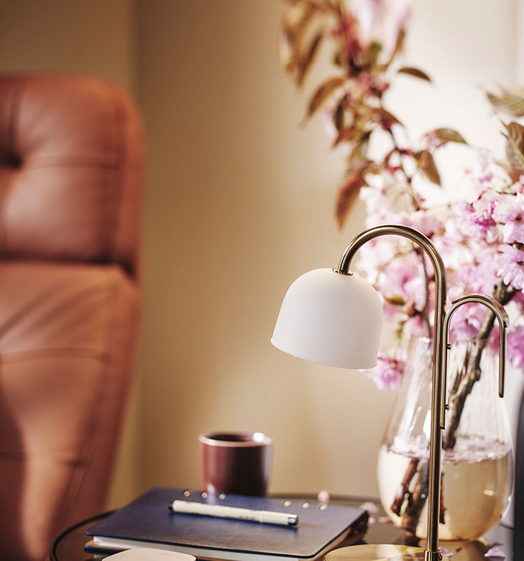 A side table with a notebook and pen, pink flowers in a vase and TVÅMASTAD LED portable lamp.