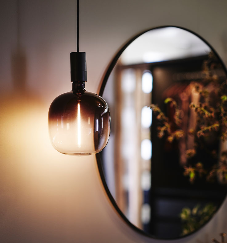 Close-up of a MOLNART light bulb with a soft light, hanging down in front of a round mirror in the background.