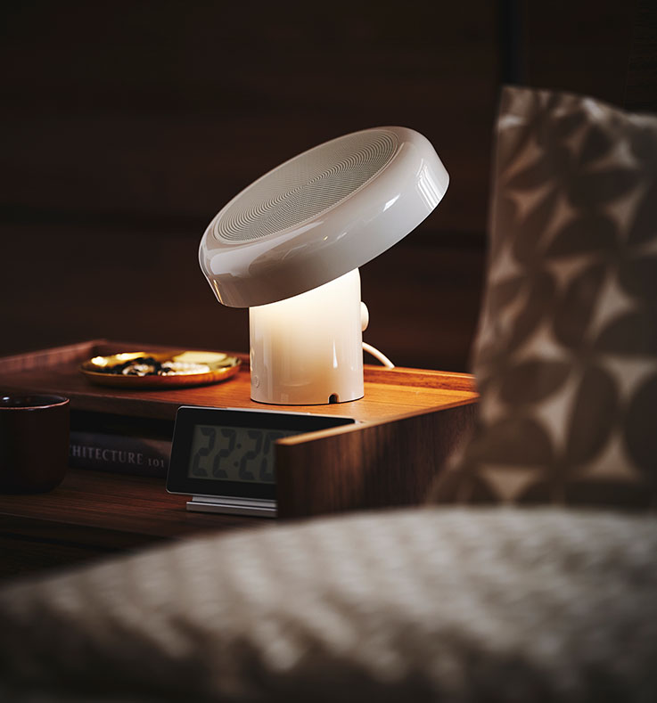 Close-up of a bedside table with BLOMPRAKT bluetooth speaker lamp, being the only light source in the otherwise dark room.