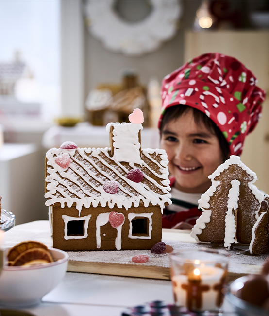 Child decorating gingerbread house with white icing and colorful candies in festive kitchen setting with candles and holiday treats