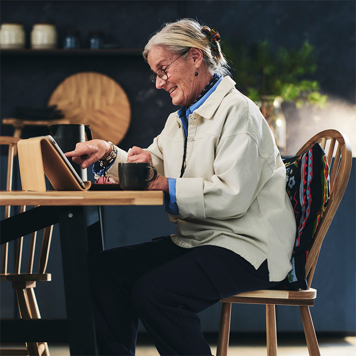 an elderly lady sitting, enjoying a coffee and looking at her tablet at the same time, at the table