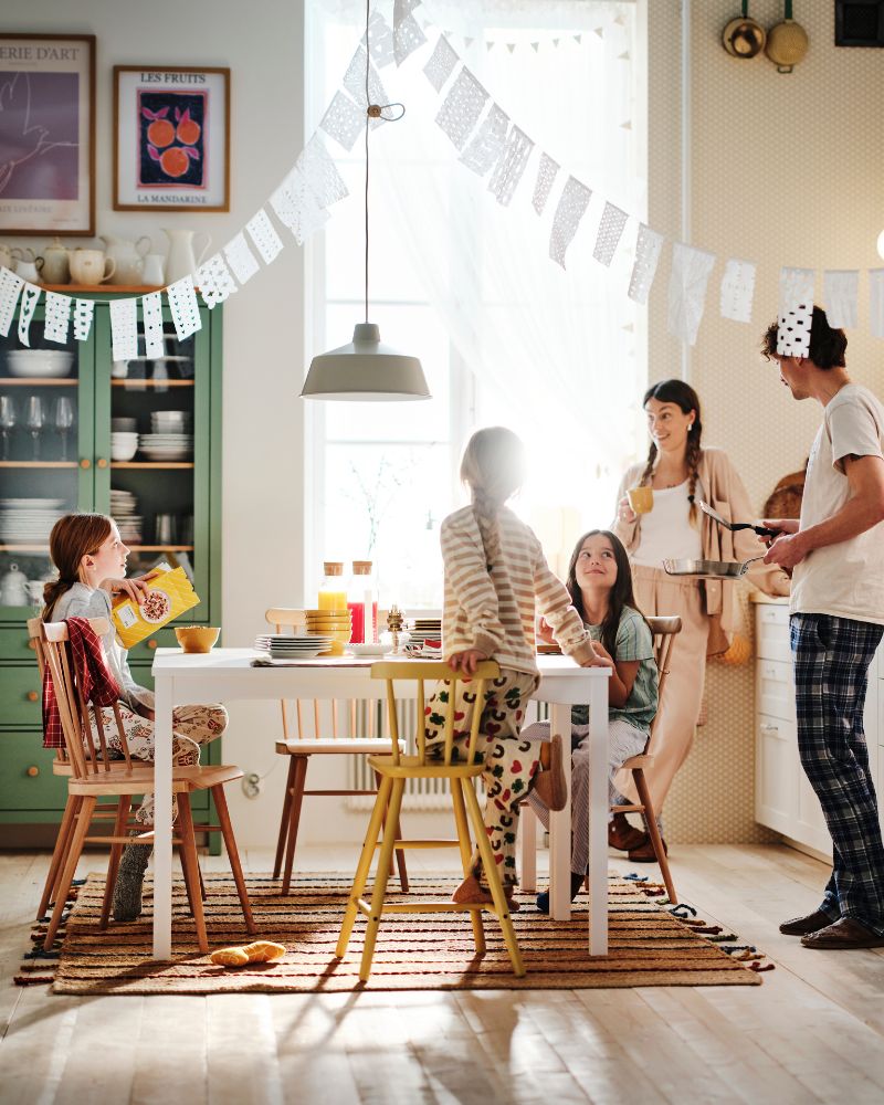 A mum and dad stand in the kitchen looking at the table where their three children are sitting enjoying breakfast.