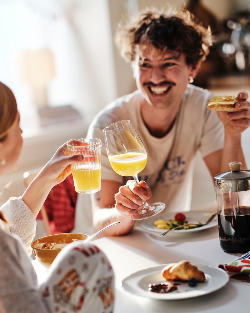 A smiling dad and his teenaged daughter sitting at the breakfast table clink their glasses of orange juice together