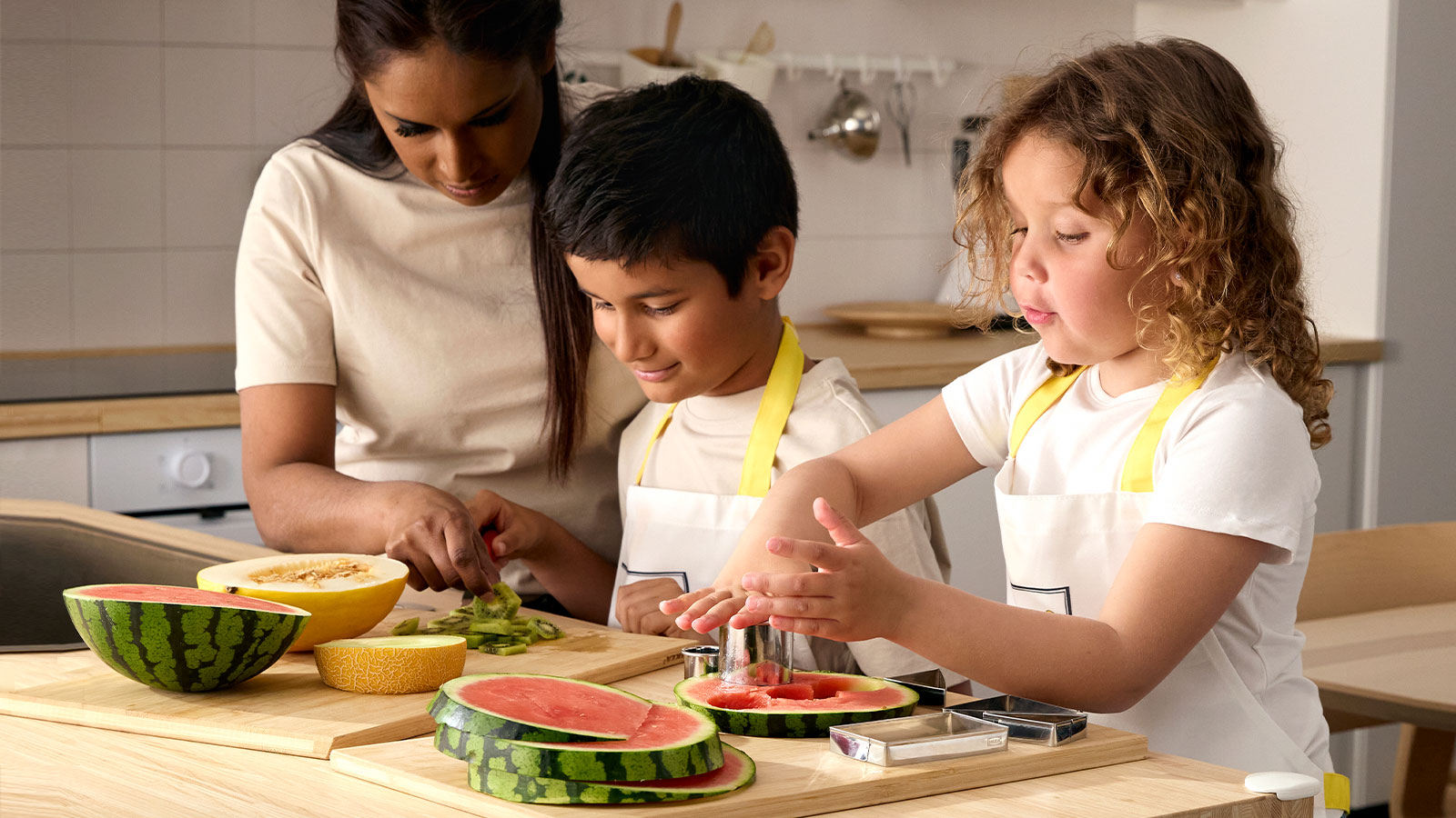 A woman and two children wearing aprons, standing by a kitchen counter cutting melon and watermelon in pieces.