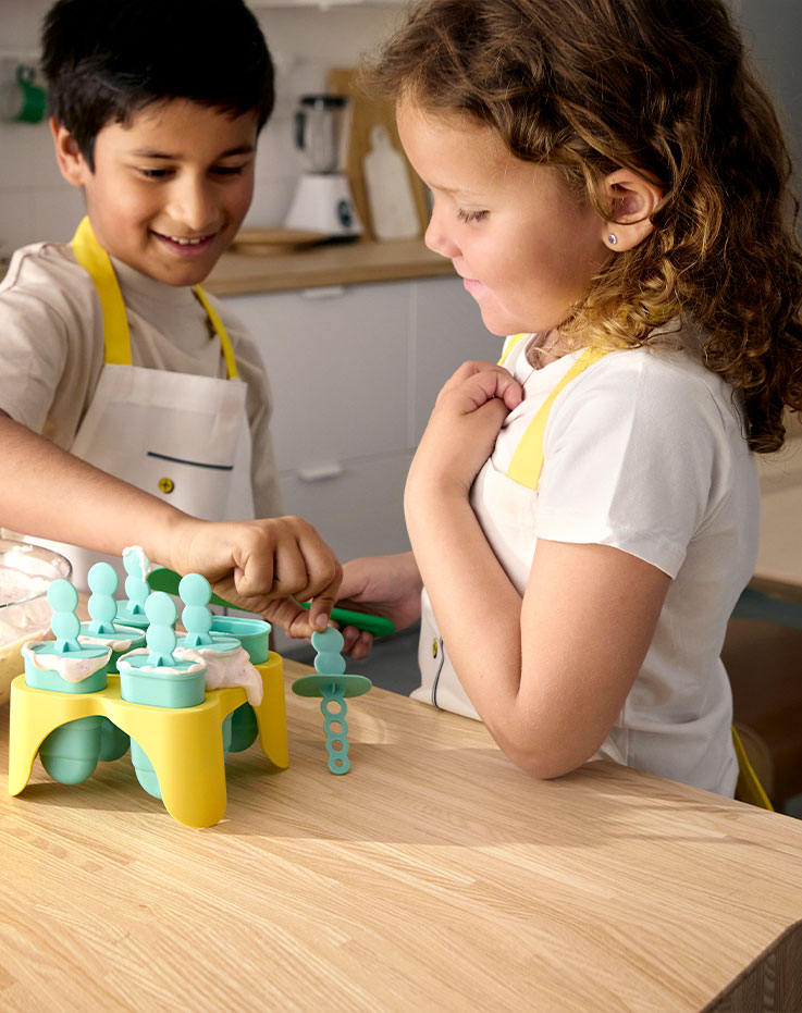 A boy and a girl standing by a kitchen counter making their own ice-cream with the help of UPPFYLLD ice lolly makers.